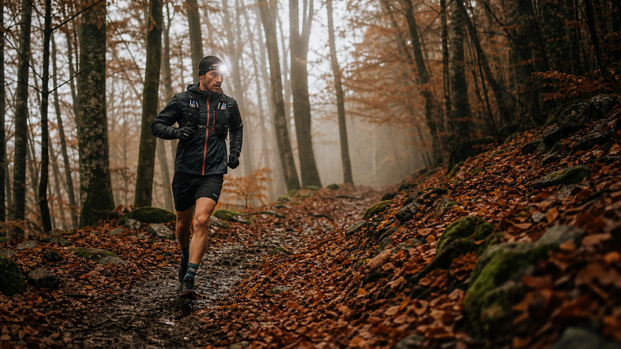 Coureur de trail sur un sentier boueux en forêt d'automne, feuilles colorées au sol et brume entre les arbres